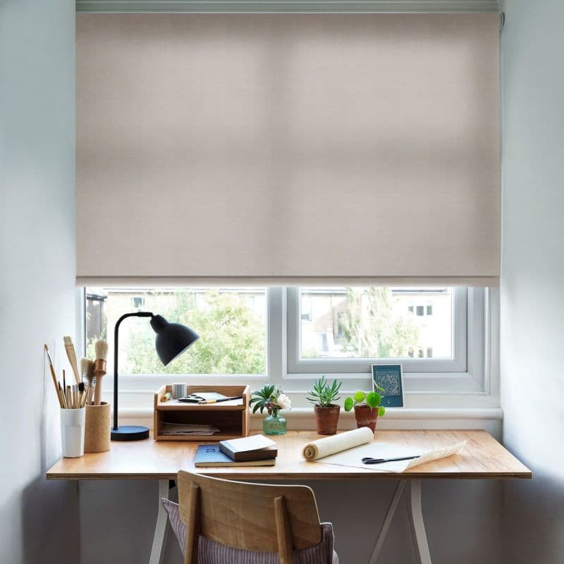 Beige roller blind, lowered nearly fully across the window, translucent fabric softening daylight; covering a small double-pane window above a wooden desk with lamp, potted plants and notebooks.