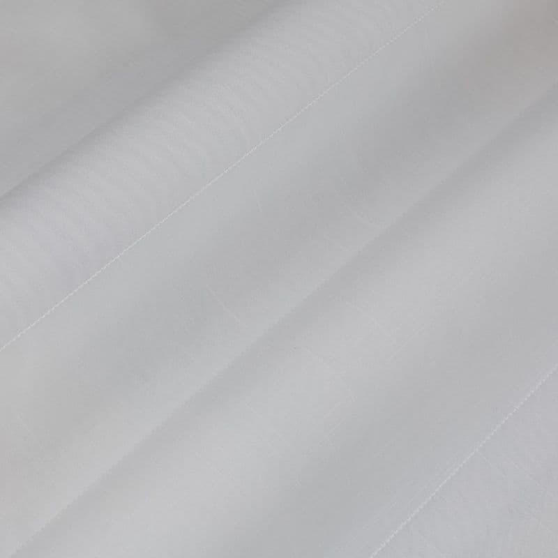 Object: white semi-sheer fabric (curtain-like) with stitched hem and faint grid texture. Action: layered into broad diagonal folds. Context: close-up view, soft diffuse lighting, neutral background.