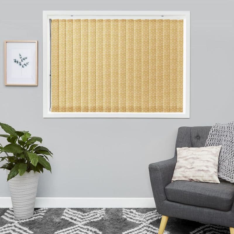 Patterned beige vertical blinds cover a window, fully closed and filtering soft light into a light-gray living room with a potted plant, framed print, gray armchair, and patterned rug.