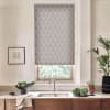 Patterned roller blind covers window above sink; natural light filters through. Kitchen countertop holds faucet and double sink, bowl of greens, potted plant, towel, and framed sketch on wall.