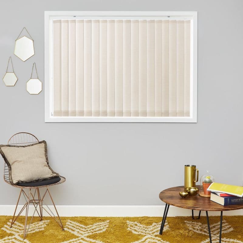 Beige vertical blinds, fully closed across a white-framed window. They filter soft daylight. Context: gray wall with three hanging hexagonal mirrors, wire chair with cushion, round wooden coffee table.
