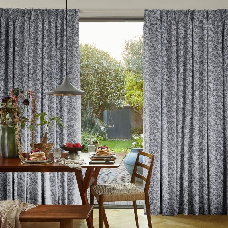 Floor-length grey curtains with dark leaf print, pleated and drawn aside; filtering daylight through sliding glass door. Dining table set for breakfast under pendant lamp, view of sunlit garden.