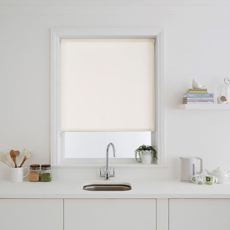 Cream fabric roller blind fully lowered over a recessed window, softly diffusing light — above a white kitchen sink with stainless faucet, minimal countertop, jars, potted plant, kettle.