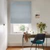 Pale blue Roman blind, lowered in soft folds, filtering daylight through a narrow bottom gap; desk beneath with mirror, vase of flowers and a teal-upholstered chair in a neutral study.