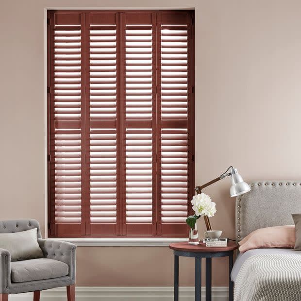 Wooden plantation shutters cover a tall recessed window—four-panel, mahogany louvers—mostly closed with slats angled to admit thin daylight; bedroom setting with upholstered bed, bedside table, lamp and armchair.