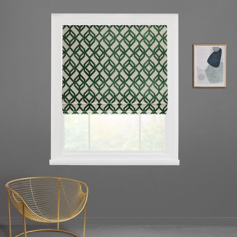 Roman blind with green geometric lattice, lowered to mid-window and filtering daylight; installed in a white frame on a gray wall, above a gold wire chair and a small abstract framed print.