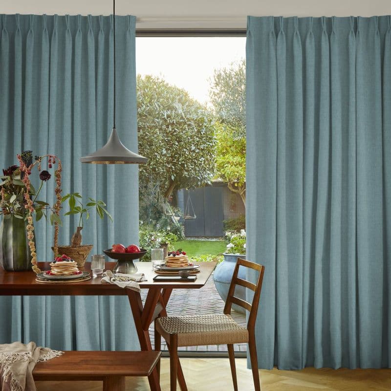 Teal pleated floor‑length curtains, drawn aside to frame a central sliding door; filtering soft daylight. Dining table with stacked pancakes and pendant lamp in dining area overlooking a leafy backyard.
