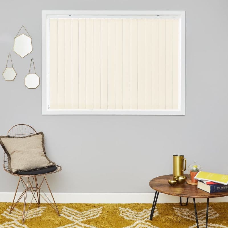 Vertical cream-colored blinds covering a rectangular window, closed and gently filtering daylight; set on a grey living-room wall with a wire chair, hanging mirrors, wooden coffee table and mustard rug.