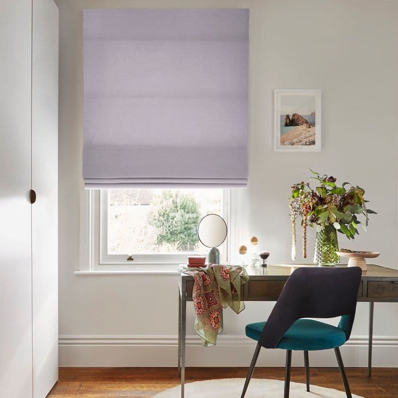 Lilac Roman blind, lowered to the sill with soft horizontal folds, filtering daylight; above desk with teal chair, mirror, vase of foliage and framed coastal print in bright study.
