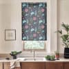 Floral roller blind covers a window, displaying teal and pink blooms; light filters beneath. Stainless faucet and sink below, countertop with bowls of greens, framed sketch left, tall plant right.