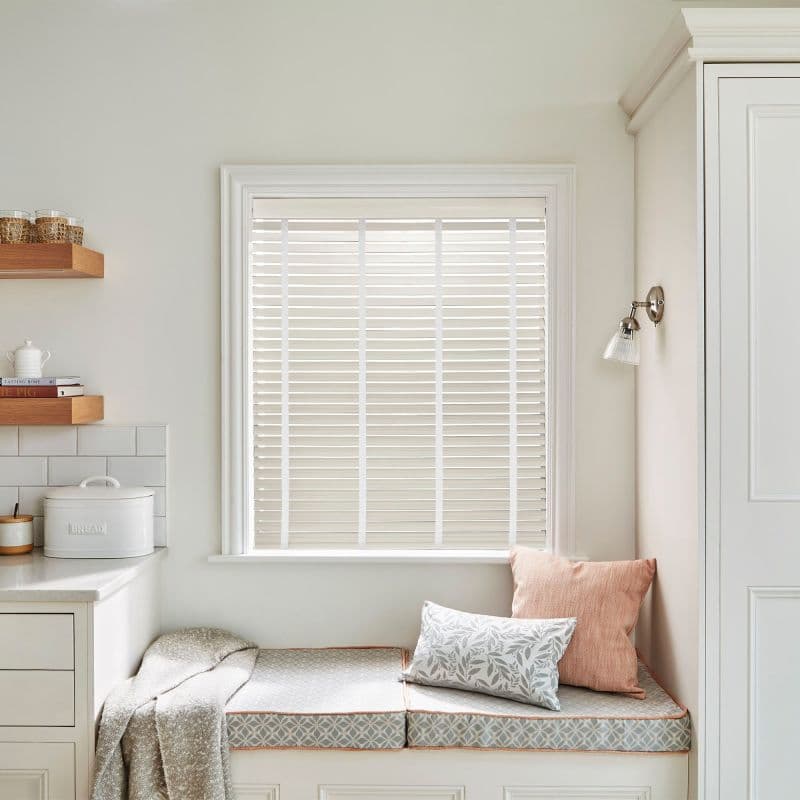 White horizontal venetian blinds covering the window, fully lowered with narrow slats filtering soft daylight; kitchen nook with built-in bench, patterned cushions, throw, wooden shelves, bread box, sconce.
BREAD
