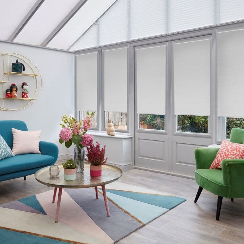 White fabric roller blinds lowered over tall windows and French doors, filtering daylight; modern sunroom with teal sofa, green armchair, round glass-topped coffee table, pastel rug and vases of flowers.