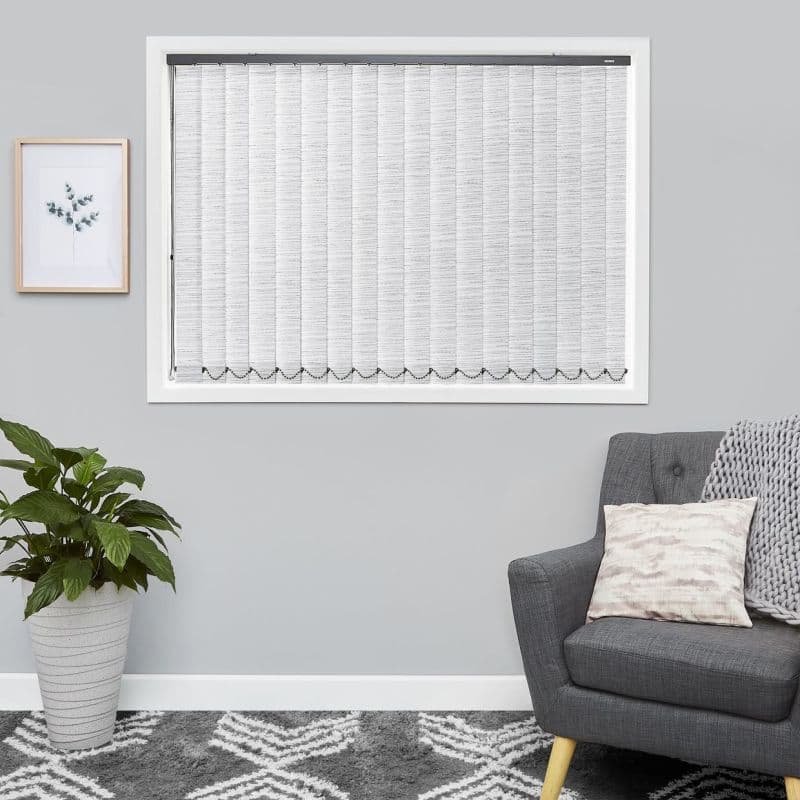 Vertical fabric blinds spanning a framed window, closed and light-filtering with scalloped bottom trim; set in a grey-walled living room with a grey armchair, throw, patterned rug and potted plant.