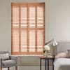 Wooden plantation shutters covering a tall window, closed with angled slats filtering warm daylight; set in a neutral bedroom corner with upholstered bed, bedside table, reading lamp and armchair.