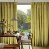 Full-length pleated mustard-yellow curtains, textured and floor-to-ceiling, are parted centrally to frame a glass door and filter daylight, revealing a dining table with breakfast settings, pendant light, sunlit garden.