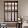 Dark wooden plantation shutters, four tall panels with adjustable louvers, closed and filtering daylight; set within a recessed window in a neutral-toned bedroom beside an upholstered bed and armchair.