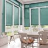 Teal horizontal Venetian blinds covering tall paneled windows and glass doors, partially lowered and filtering soft daylight. Context: bright sunlit dining nook with white table, chairs, and garden view.
