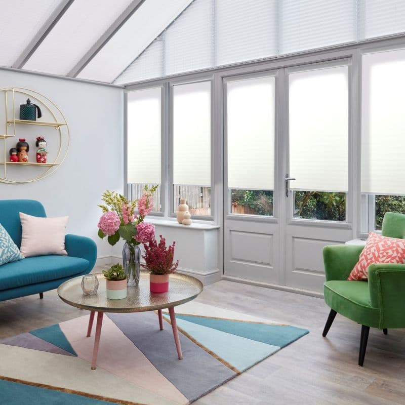 White pleated cellular blinds covering tall glazed windows and door, pulled down to diffuse daylight with lower edges slightly raised; bright conservatory-style living room with colorful sofas and garden view.