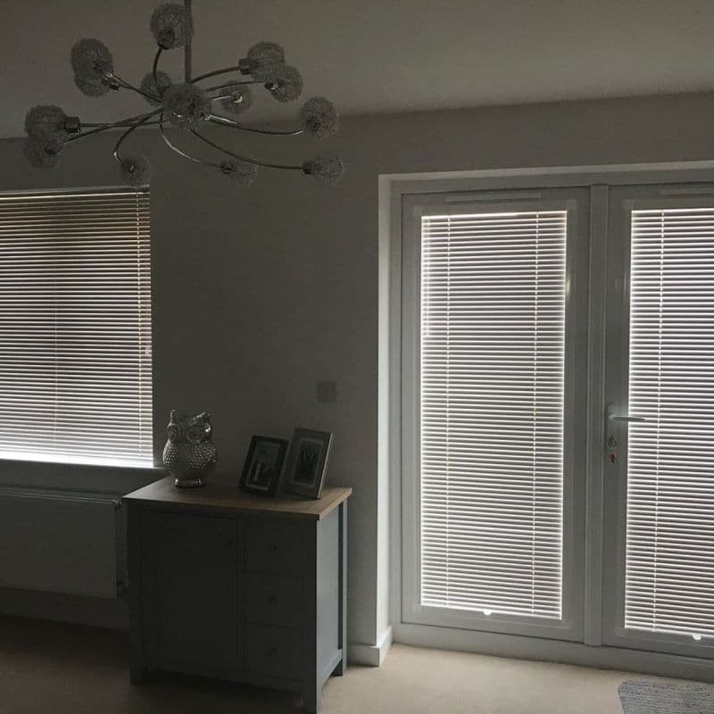 Horizontal venetian blinds on a window and patio door, closed and filtering dim daylight; living-room corner with a small cabinet, owl ornament, two photos, and chandelier.