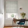 Roman blind with pale botanical pattern, lowered in folds over the window, filtering daylight; above a desk with mirror, teal chair and vase of flowers in a lit home office.