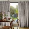 Pleated curtains with a multicolored chevron-dot pattern, hung either side of a large sliding glass door, parted to admit sunlight; dining table with breakfast setting overlooks a lush backyard patio.
