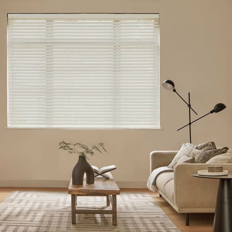 White horizontal blinds fully lowered and closed, softly filtering daylight through slats; beige living room with a neutral sofa, wooden coffee table, patterned rug and black floor lamp.