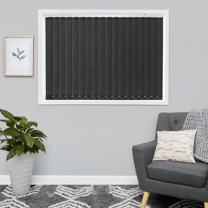 Vertical dark grey blinds cover a white-framed window, fully closed with beaded chain weights; pale grey living-room wall flanked by a potted plant left and an upholstered armchair right.