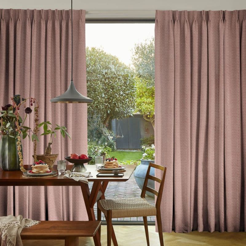 Pleated dusty-pink floor-length curtains with subtle texture, parted at the center to reveal a sunlit garden; a wooden dining table set for breakfast, pendant lamp and woven chairs.