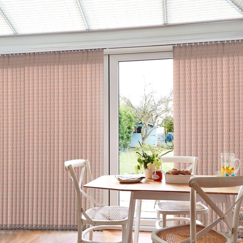 Patterned vertical curtains (pale pink geometric print) hang full-length, parted at the center to reveal glass door; filtering daylight over a dining table with cane-seat chairs and garden view.