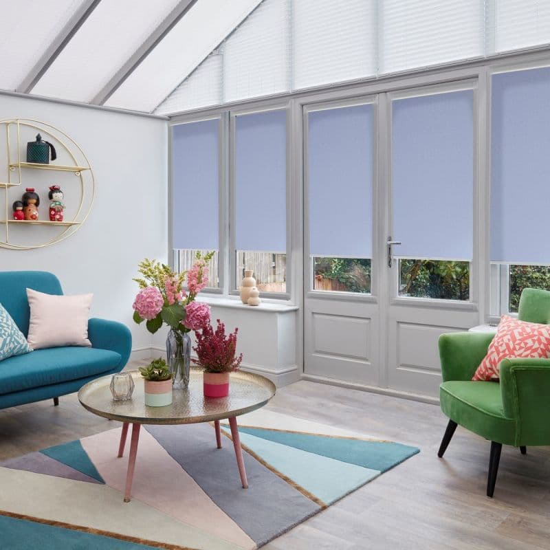 Pale blue roller blinds lowered on French doors and tall windows, with pleated roof blinds above; filtering daylight. Bright conservatory living room with teal sofa, green armchair, pastel rug.