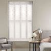 White full-height plantation shutters with four framed panels and horizontal louvers, closed and filtering bright daylight; set in a neutral-toned bedroom corner with upholstered bed, side table, lamp, and armchair.