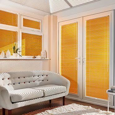 Honey-yellow horizontal Venetian blinds covering windows and French doors, closed and filtering sunlight into slatted beams, casting striped shadows across a cream sofa in a bright daytime living room.