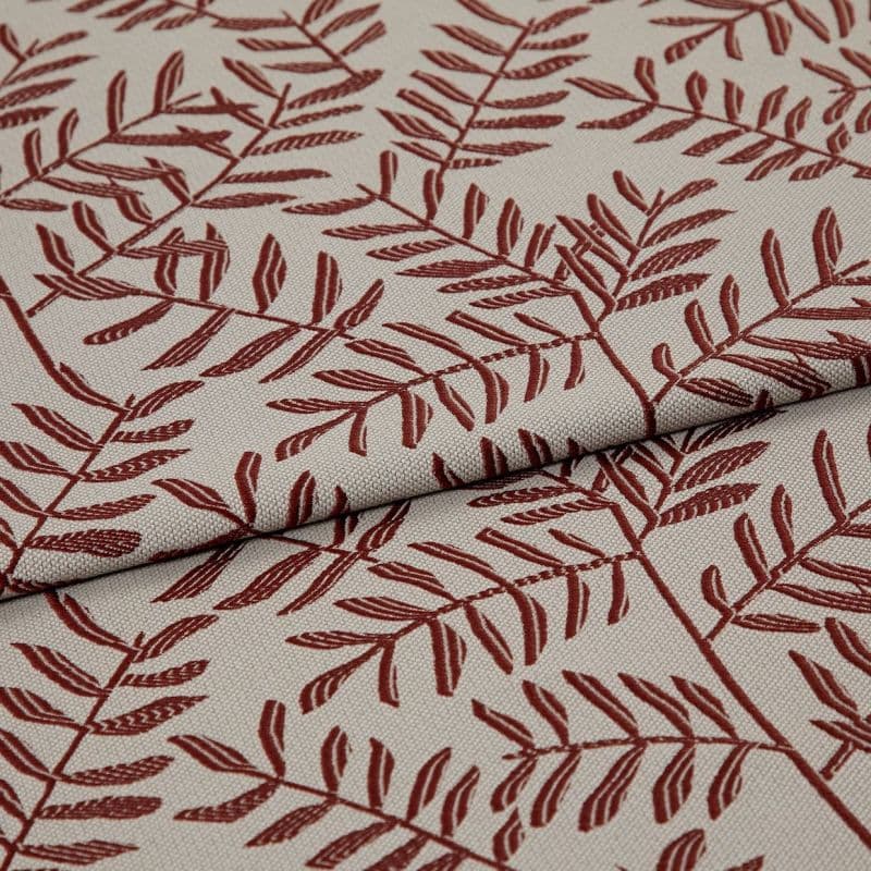 Woven beige upholstery fabric patterned with rust-red fern branches, folded and overlapped across the frame, close-up studio-lit view highlighting the woven texture and printed details.