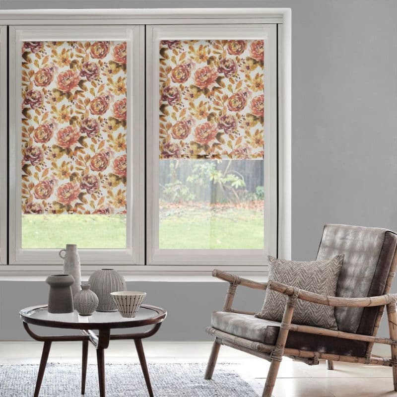 Floral roller blinds with warm rose-and-leaf pattern cover two adjacent windows, one fully lowered and one half-lowered, filtering daylight into a neutral-toned living room with chair and coffee table.