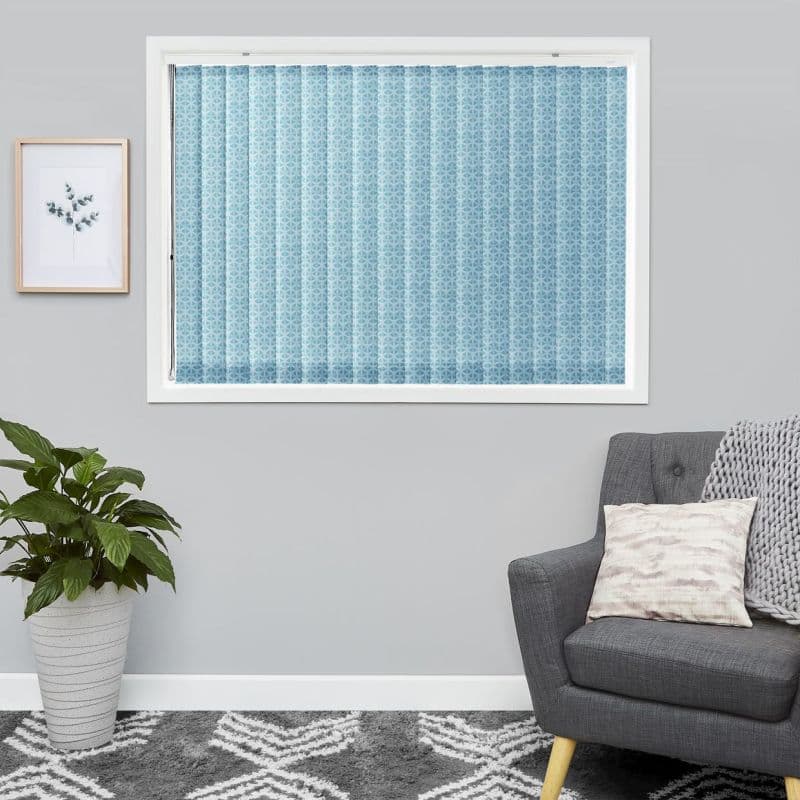 Light-blue patterned vertical blinds, fully closed and filtering light across the window; in a neutral gray living room with a potted plant, framed botanical print and gray armchair.