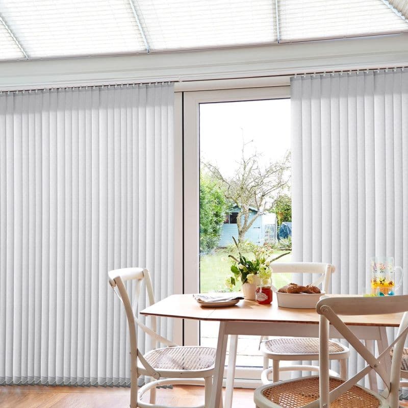Pale gray vertical blinds, full-height fabric slats on a ceiling track, partially open; filtering daylight into a dining nook with wooden table, woven-seat chairs, and visible backyard garden.