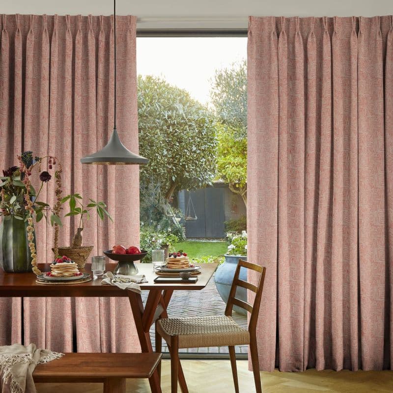 Pink pleated full-length patterned curtains, drawn to each side revealing a central glass patio door; filtering soft daylight into a dining area with wooden table, pendant lamp and garden.