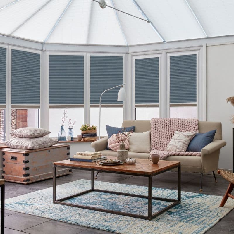 Blue pleated cellular blinds, lowered across tall conservatory windows with slim frames, filtering daylight while leaving lower panes exposed; cozy sunroom with sofa, coffee table, pale rug and diffused light.