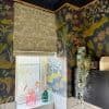 A patterned fabric Roman blind, lowered over a frosted window, filters daylight; botanical navy-and-gold wallpaper frames a tiled bathroom sill topped with glass vases and cascading faux foliage.