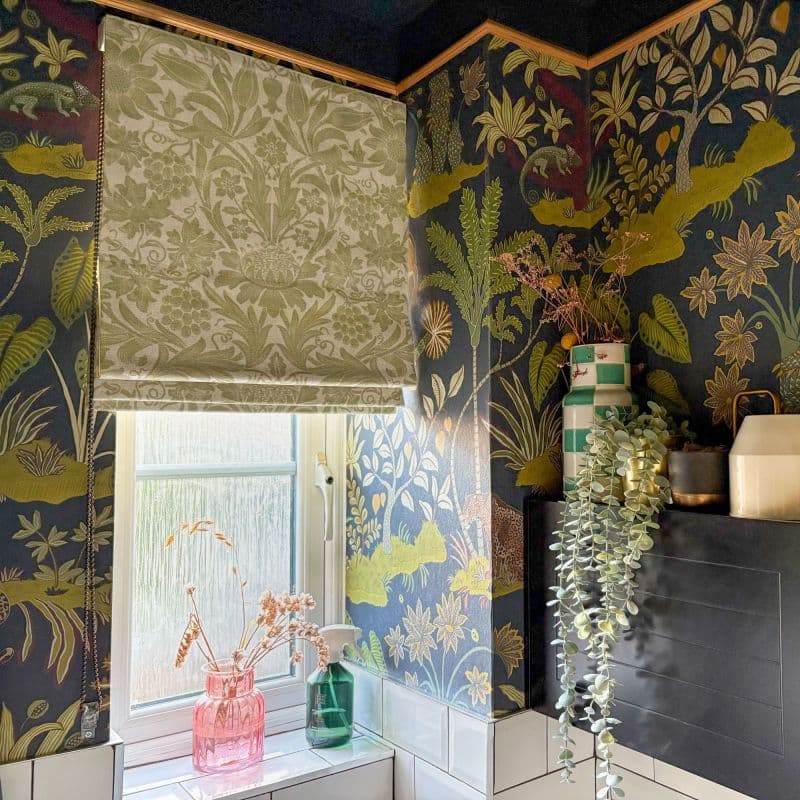 A patterned fabric Roman blind, lowered over a frosted window, filters daylight; botanical navy-and-gold wallpaper frames a tiled bathroom sill topped with glass vases and cascading faux foliage.