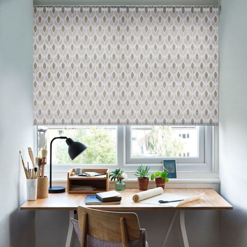 A patterned roller blind, white with beige leaf motif, is fully lowered over a double window, filtering daylight above a wooden desk with lamp, brushes, potted succulents and urban view.