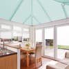 Seafoam pleated roof blinds cover the pitched conservatory ceiling; white pleated blinds cover windows and doors, filtering soft daylight into a bright kitchen-dining conservatory with wooden table and garden view.