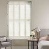 White plantation shutters covering a tall window, closed with angled louvers filtering soft daylight; bedroom setting with upholstered bed, round side table with lamp and flowers, armchair, and neutral walls.