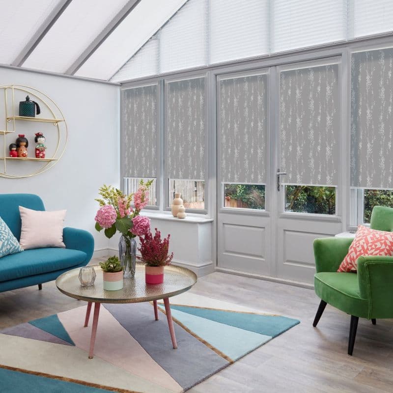 Patterned grey roller blinds on conservatory windows and door, lowered leaving small clear lower panels; filtering daylight into a bright sunroom with colorful seating and a flowered coffee table.