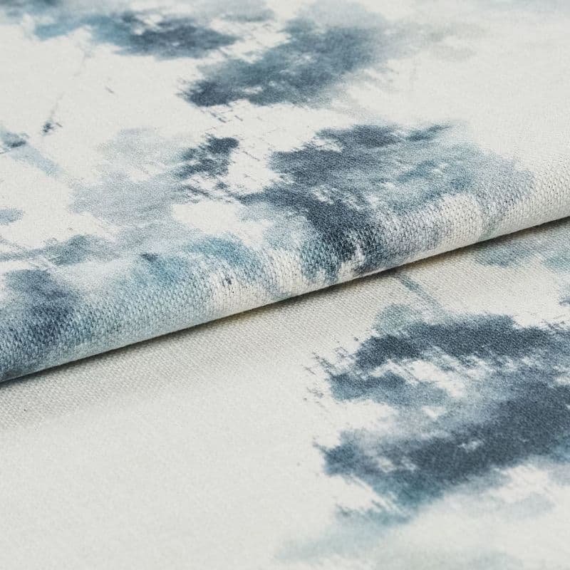 Folded woven fabric patterned with blue‑gray watercolor blotches is layered in a close-up, softly lit view, revealing coarse weave texture against a muted off‑white background.
