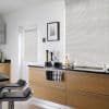 White horizontal blinds lowered over a large window and adjacent door, closed and softly filtering daylight; modern kitchen with light wood cabinets, black countertop, two bar stools and minimal decor.