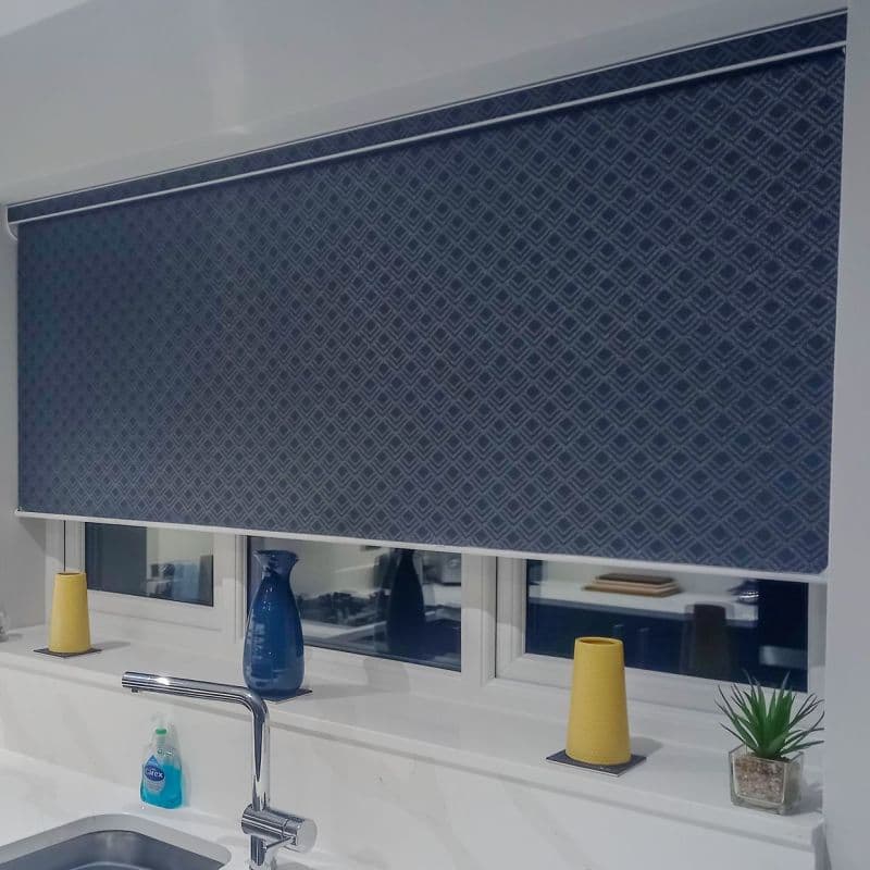 Patterned roller blind (navy diamonds) fully lowered, blocking view and filtering light; kitchen window above sink with faucet, blue vase, yellow votives, soap bottle and small potted plant on sill.
Text in image: Carex