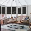 Black pleated cellular blinds lowered across conservatory windows, filtering and blocking light; in a bright living area with a beige sofa, chunky pink throw, wooden coffee table and blue patterned rug.