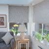 Fabric leaf-patterned Roman blinds, lowered halfway and filtering soft daylight; a sunlit bay living room with a sofa, side table and lamp, orchid and potted plants on the windowsill.