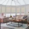Pleated honeycomb blinds covering conservatory’s tall, multi-panel windows, lowered to mid-height and filtering soft daylight; beige blinds frame a cozy sunroom with sofa, knitted throw, wooden coffee table and rug.